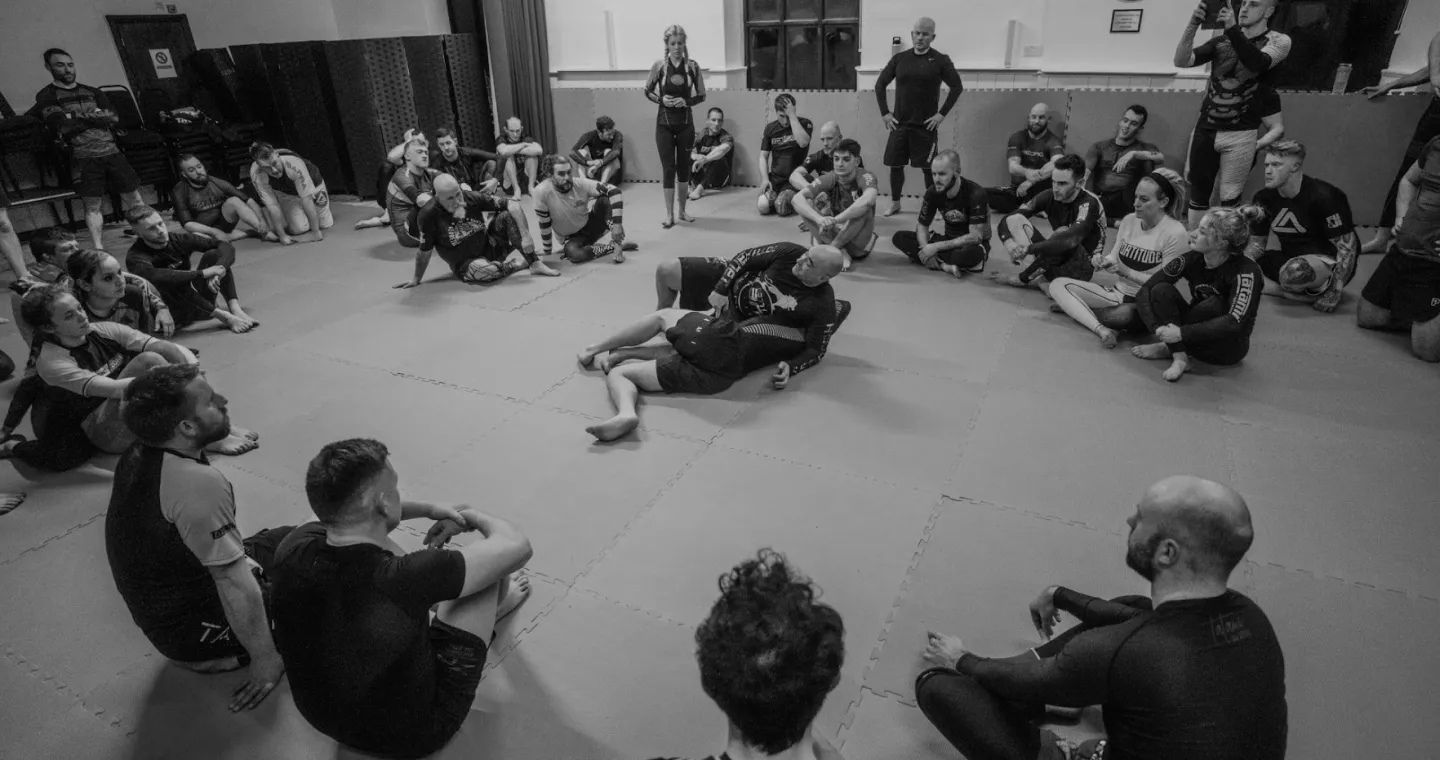 Students gathered round watching a Jiu-Jitsu coach as they demonstrate how to execute a new BJJ technique.